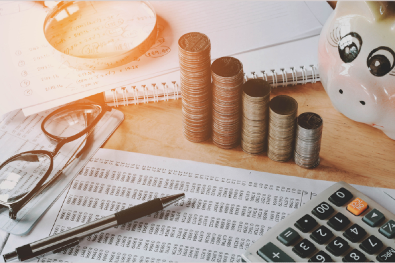 Financial reports, a calculator, pen, reading glasses, stacks of coins, a piggy bank and a magnifying glass on a desk repres