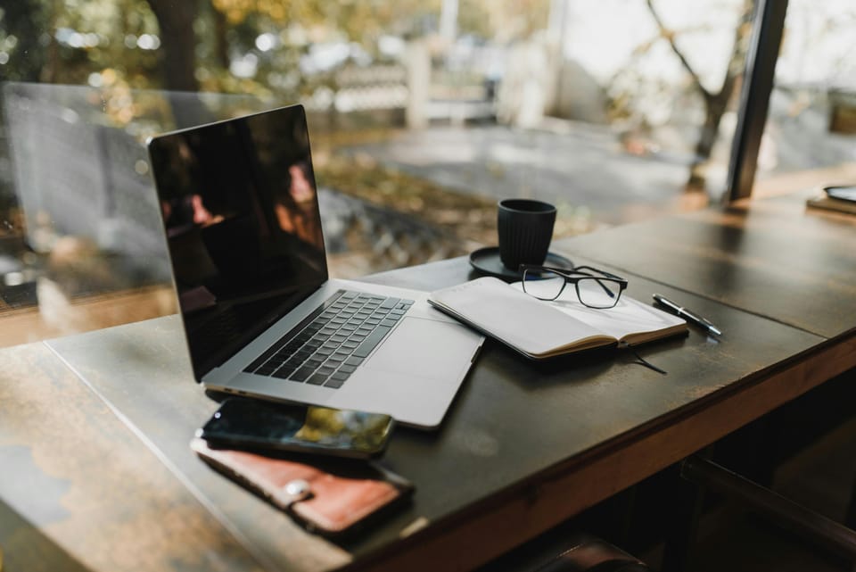 Laptop, notebook, and coffee cup on wooden desk with natural lighting, showing organized workspace setup